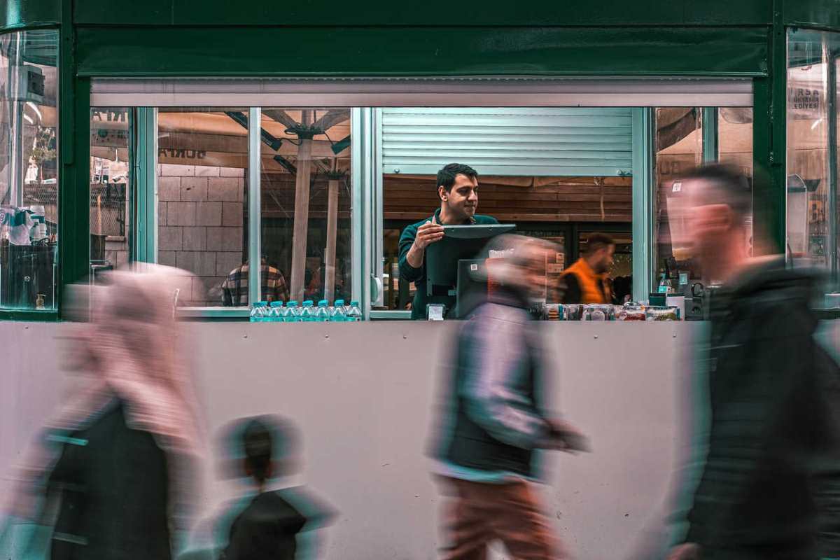 A street vendor serving customers through a kiosk window while people walk by in a busy market setting.