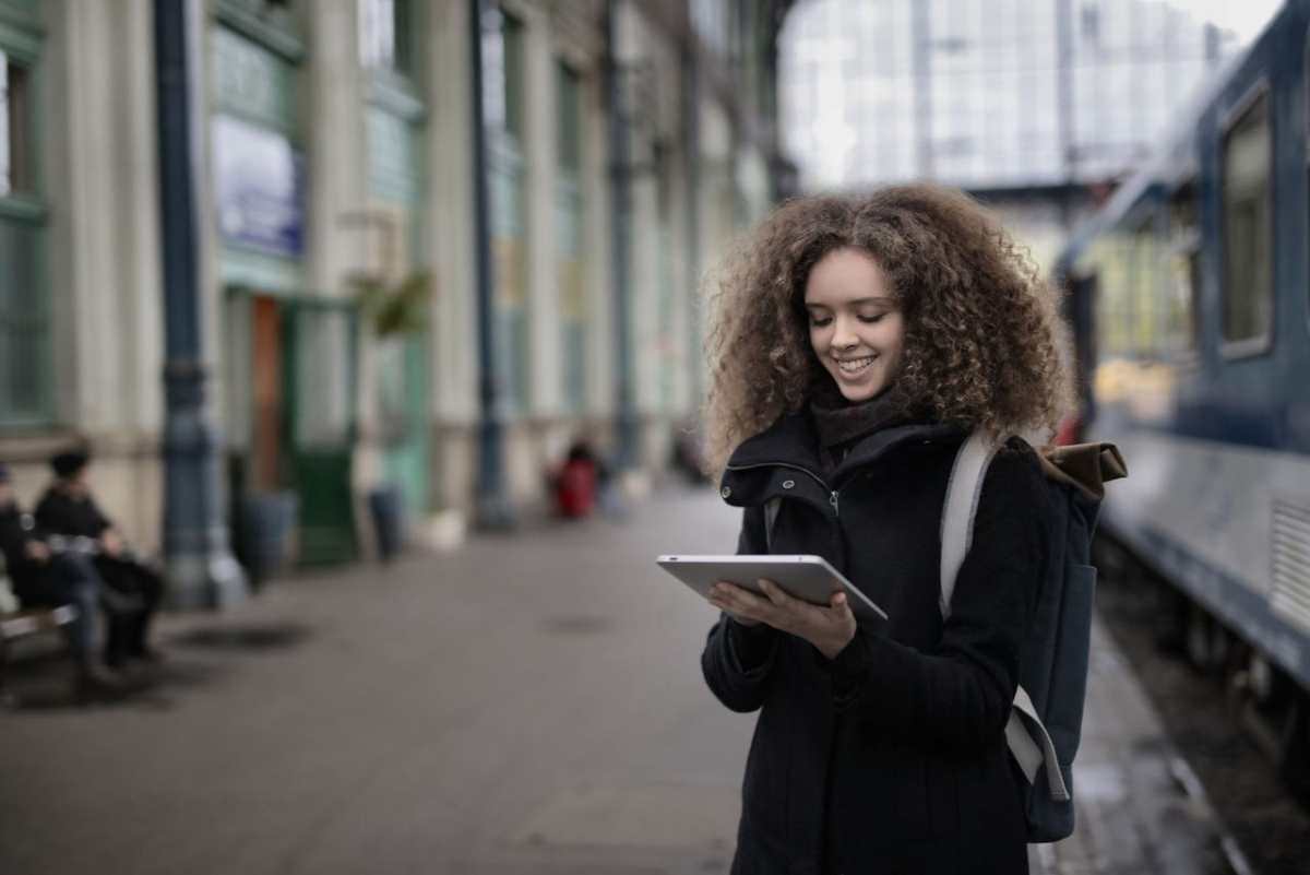 Positive young lady with curly hair in casual warm coat browsing tablet while standing on platform and waiting for train