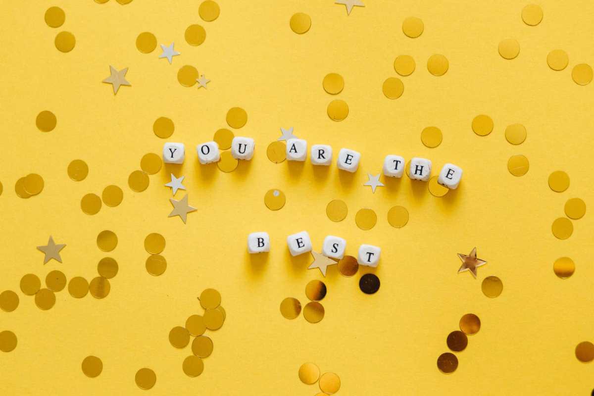 Inspirational message ‘You Are the Best’ spelled with dice on a vibrant yellow surface with star confetti.