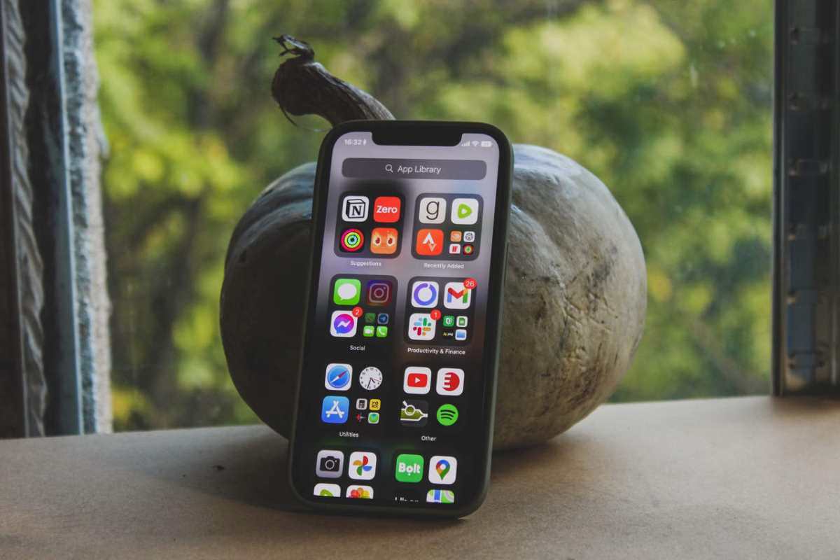A smartphone displaying various app icons stands beside a pumpkin by a window with a green outdoor view.