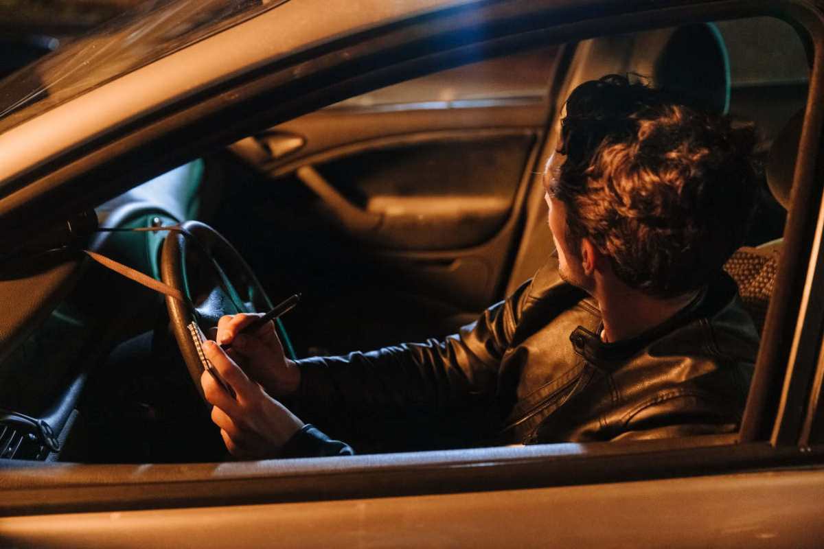 A man in a leather jacket taking notes inside a car at night, implying a stakeout or investigation setting.