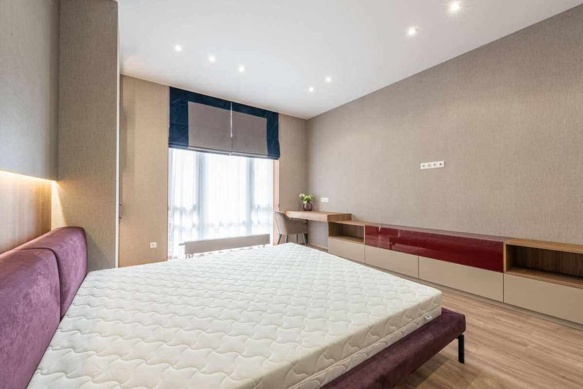 Modern bedroom with white mattress on bed placed against wooden cabinet and table with flowerpot at wall with roller blind on window
