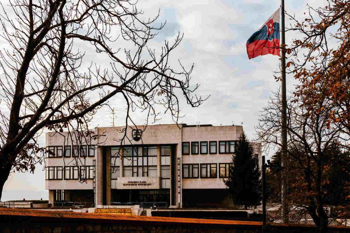 A view of the Slovak Parliament building surrounded by autumn trees with the national flag waving under an overcast sky.