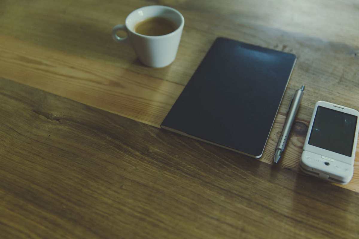 A simple workspace setup featuring a coffee cup, notebook, pen, and smartphone on a wooden desk.