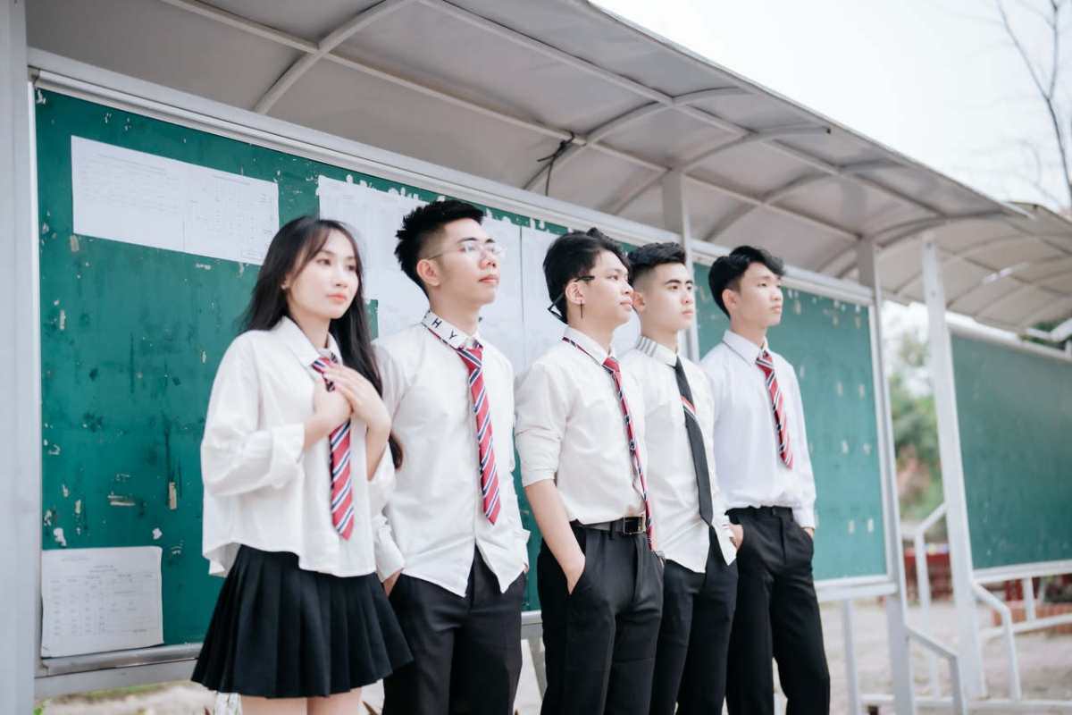 A group of five students in school uniforms standing in front of a notice board outdoors.