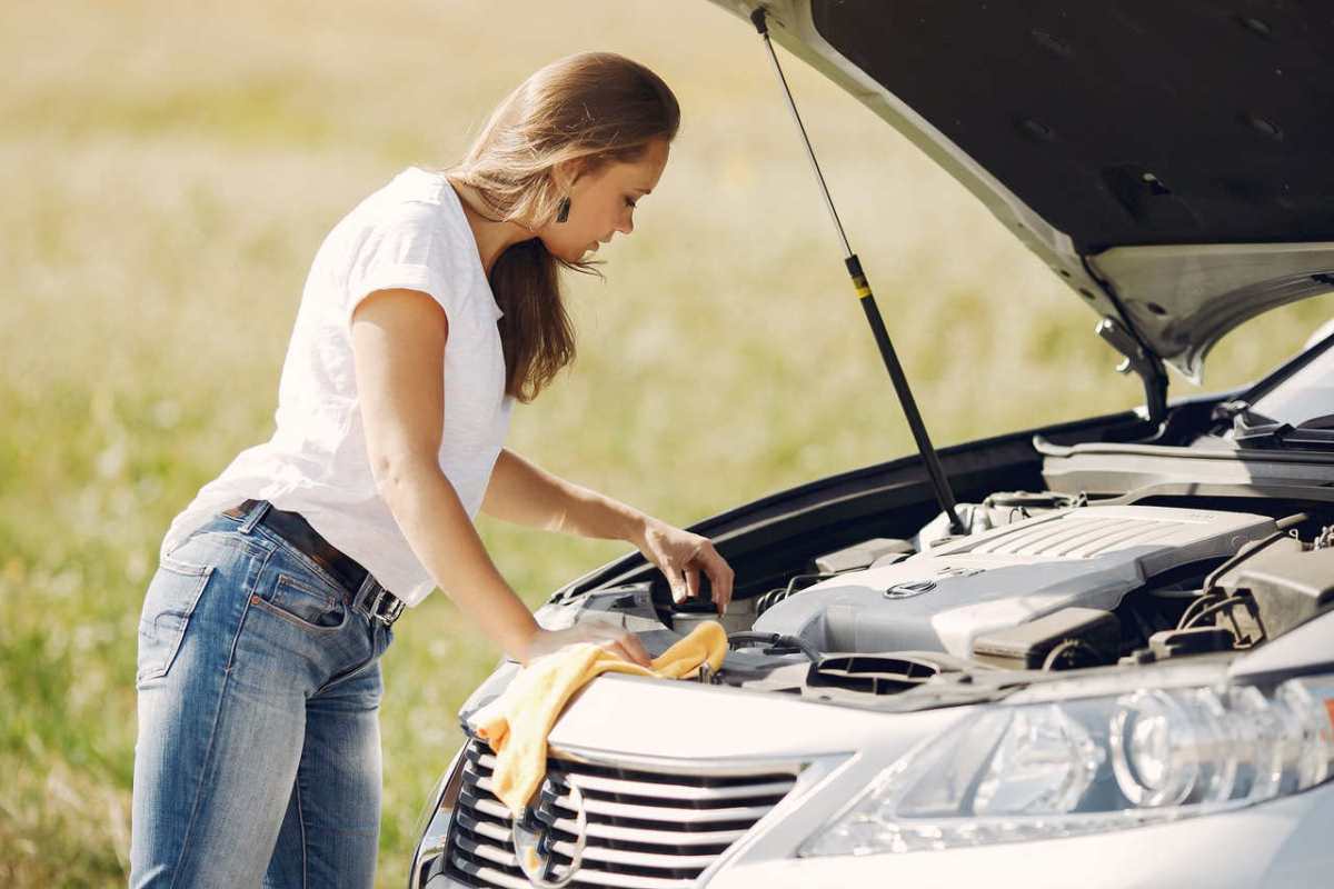 Side view of female driver in white t shirt and jeans checking damage of car engine and wiping details under open hood while standing against green field in summer day