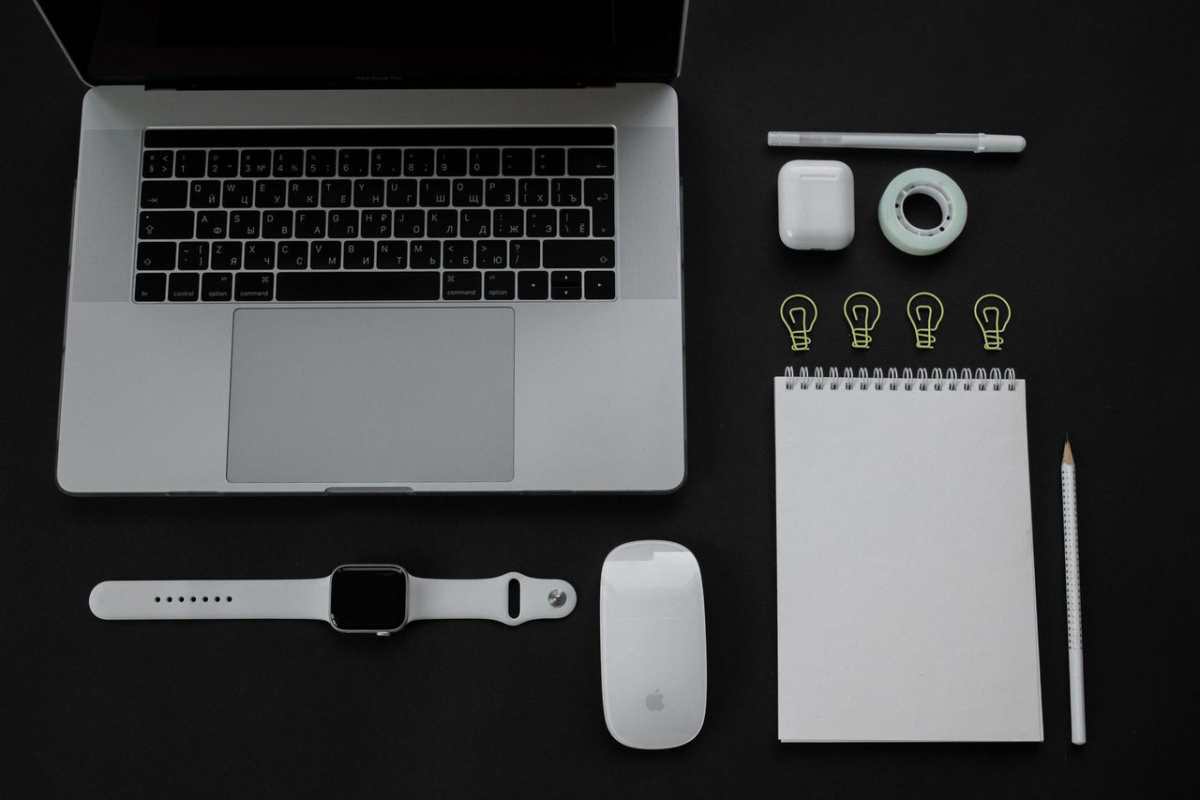 Flat lay of modern office essentials including a laptop, smartwatch, and stationery on a black background.