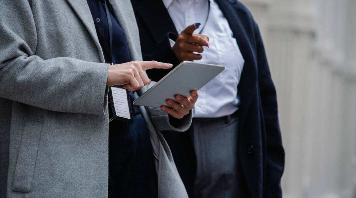 Crop unrecognizable diverse female managers in elegant coats pointing on tablet while discussing business project on street