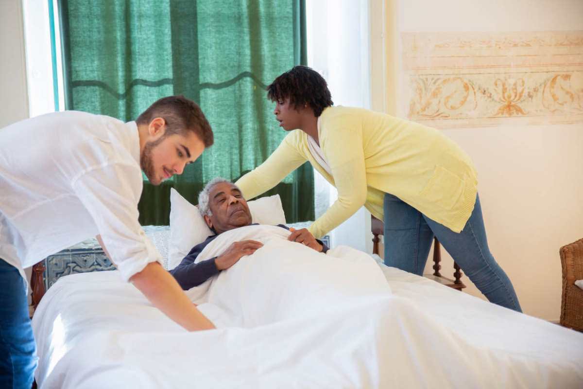Caregivers supporting an elderly man lying in bed, providing assistance and comfort.