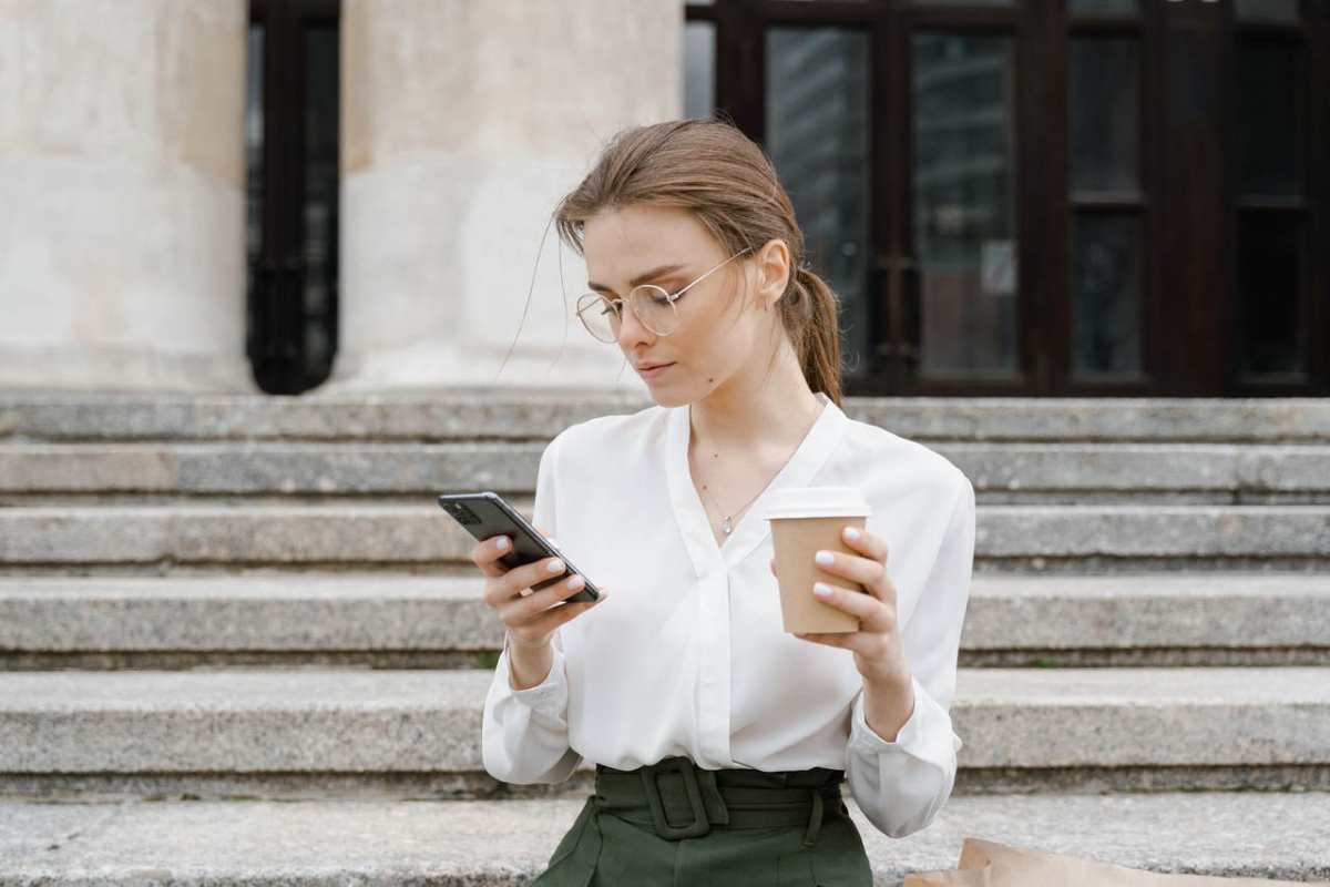 Young woman in business attire using smartphone while enjoying a coffee outdoors.